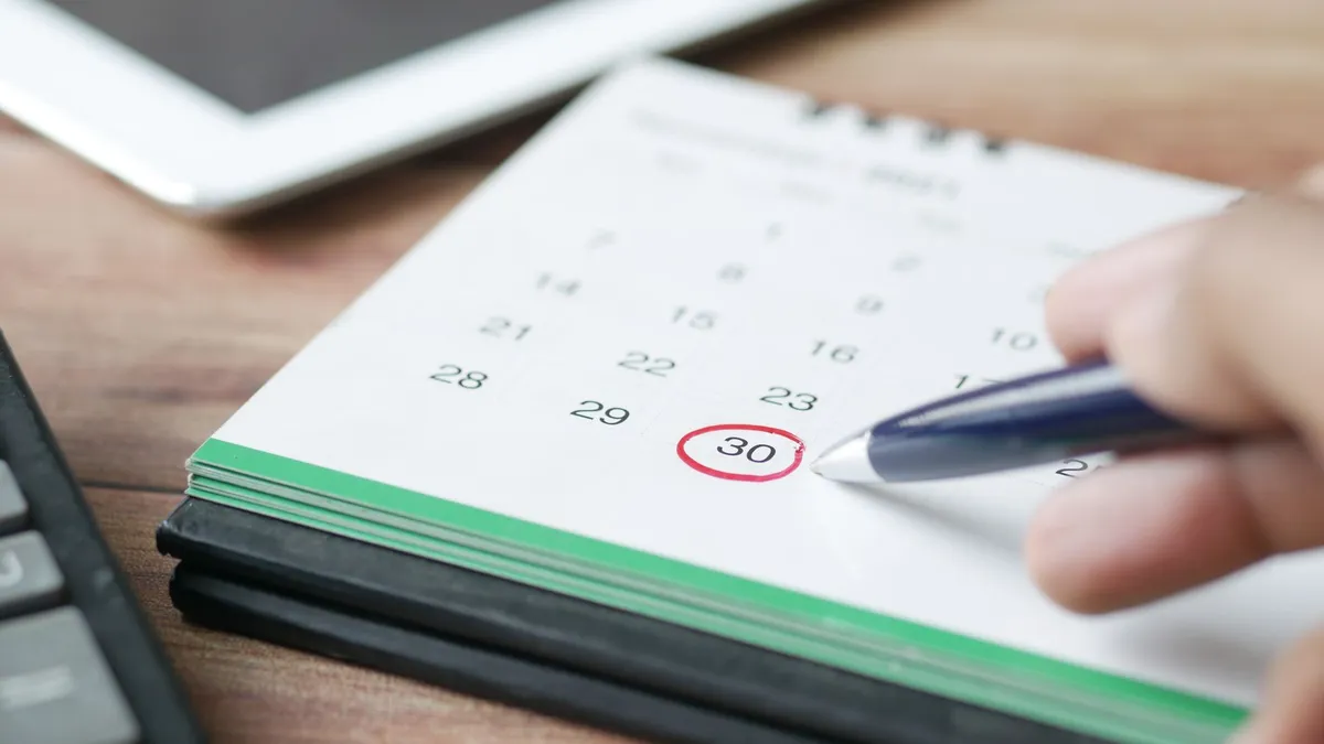 Mano marcando con rotulador rojo una fecha de plazo en un calendario de papel, sobre un escritorio con tableta y calculadora