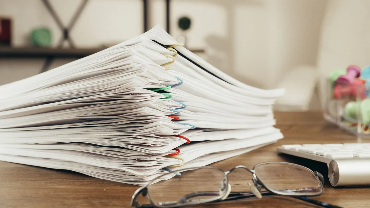 Pila de expedientes de distintos clientes separados con clips de colores sobre el escritorio de un despacho, con unas gafas y un teclado al lado